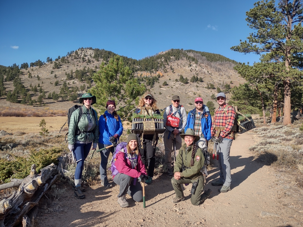 Group of eight hikers posing on a dirt trail in a mountainous area with trees and clear sky. They are wearing outdoor gear and carrying backpacks, hand tools, and seedling trees..