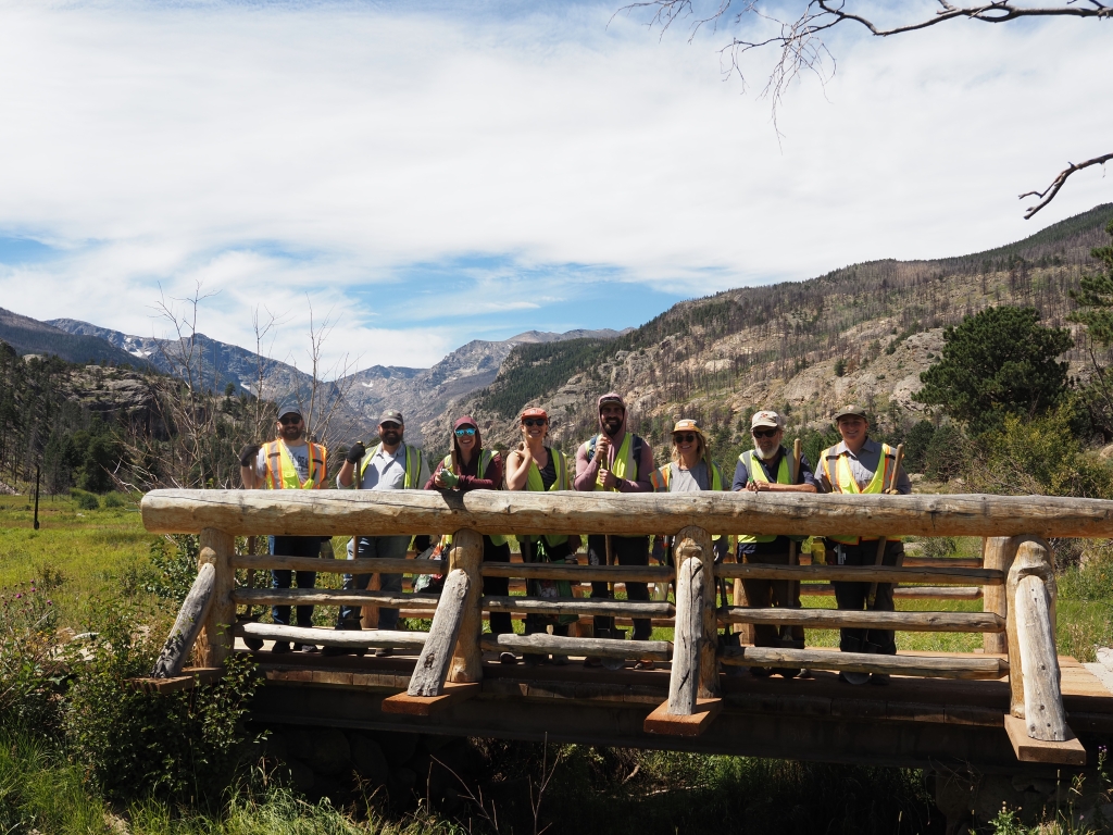 A group of people in safety vests stand on a wooden bridge in a mountainous landscape with grassy fields and sparse trees.