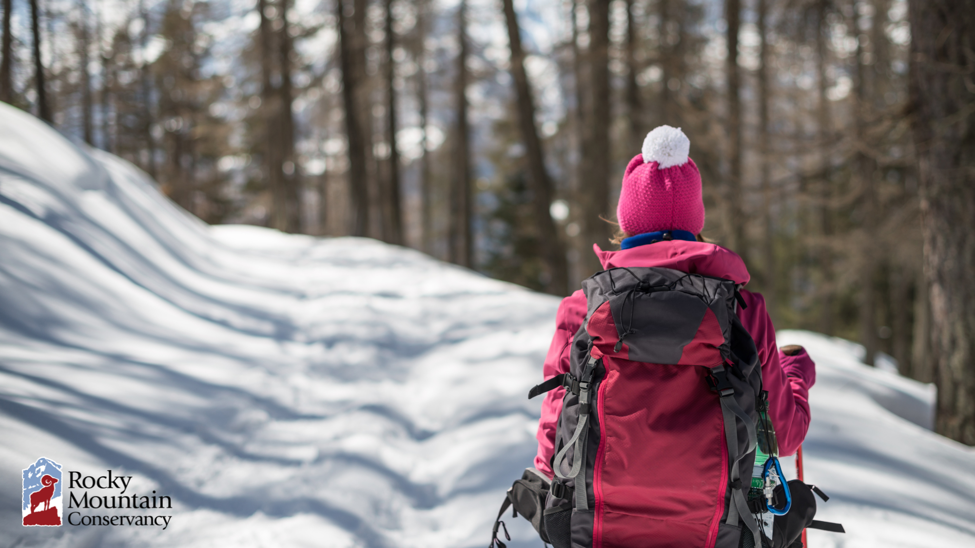 Person in pink winter attire hiking through a snowy forest, with a large backpack and walking poles. Logo of Rocky Mountain Conservancy is visible in the corner.