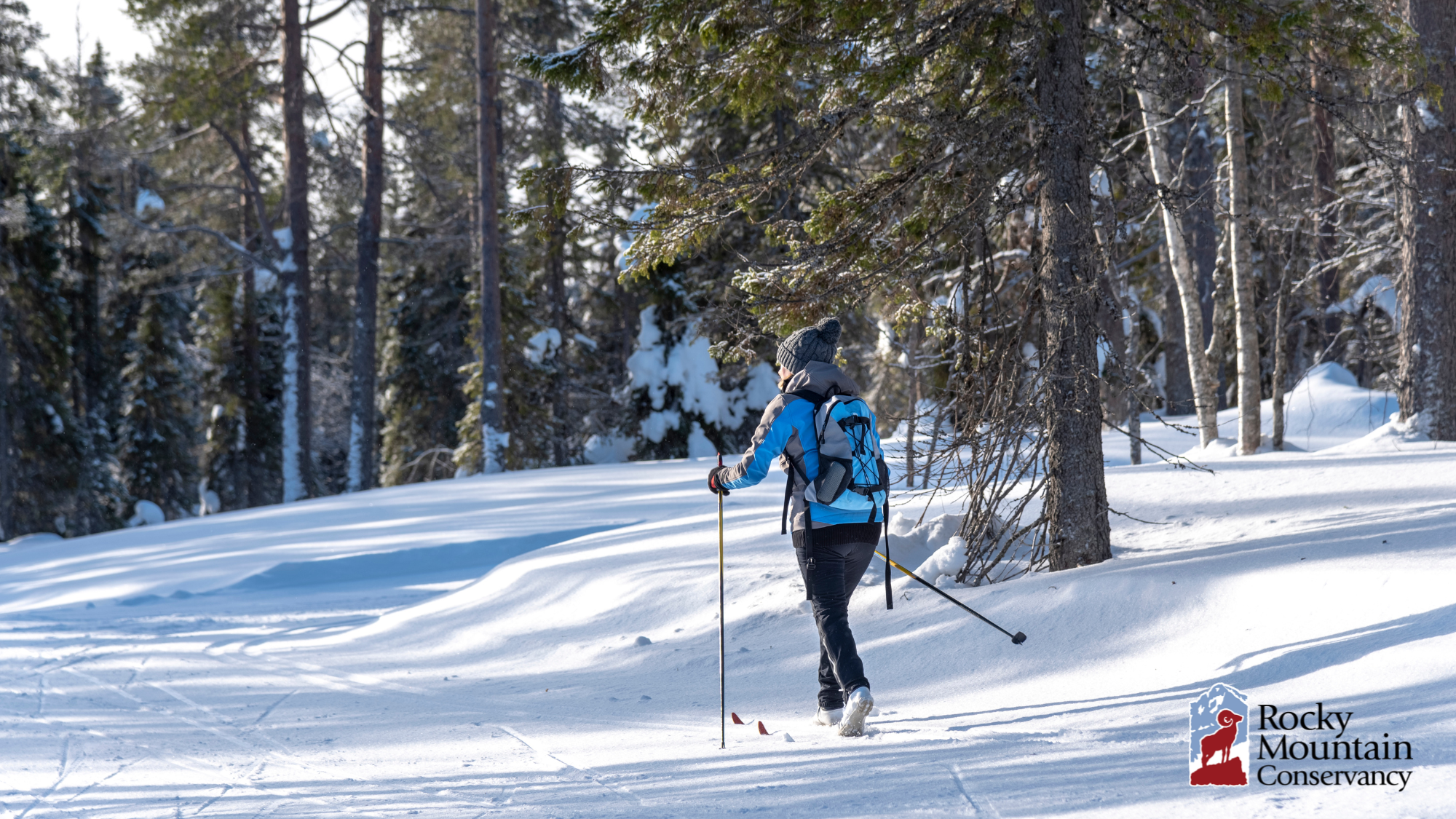 A person cross-country skiing on a snow-covered trail through a forest, carrying poles, with a blue backpack and jacket. Rocky Mountain Conservancy logo in the corner.