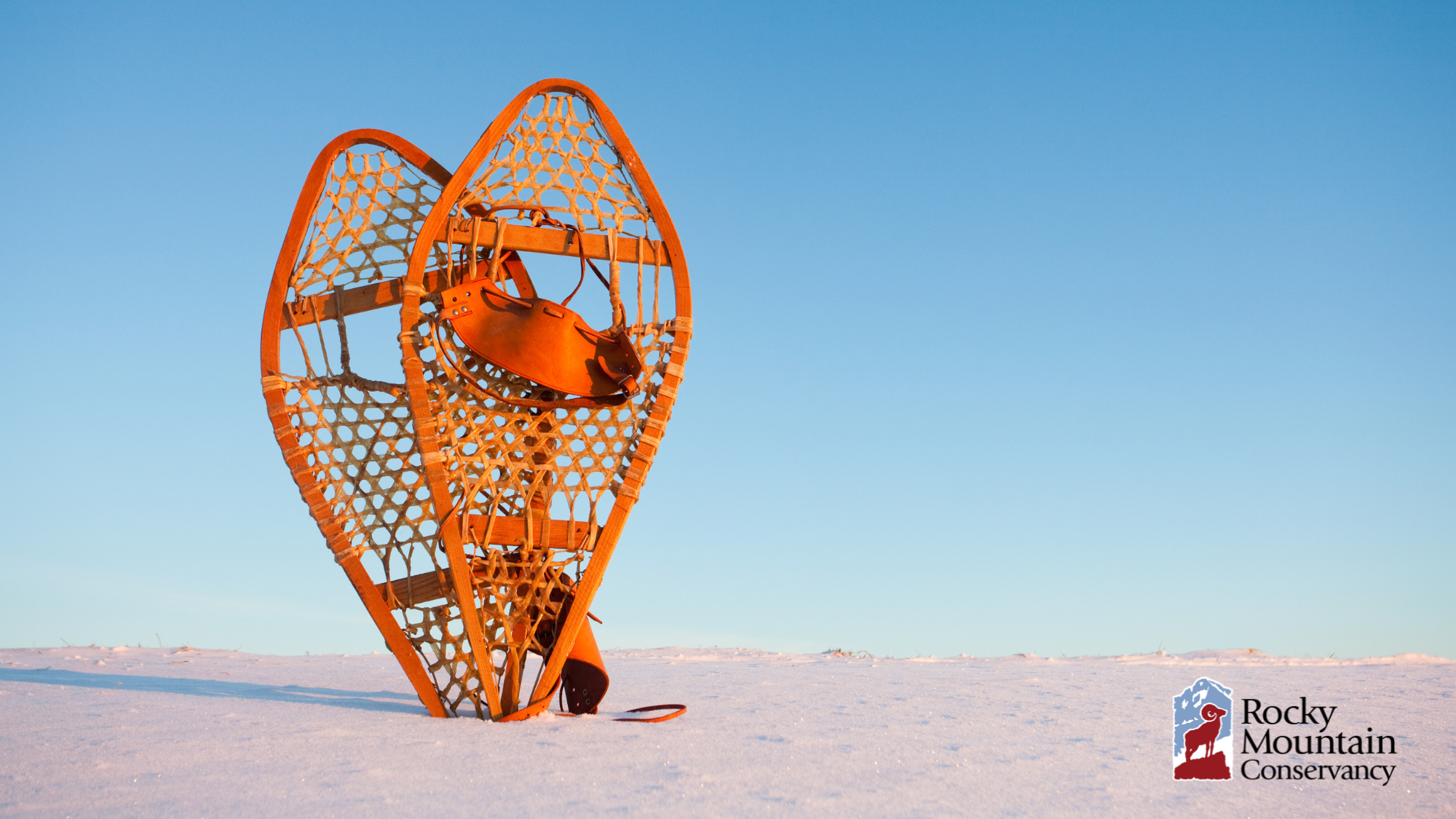 Two wooden snowshoes with leather bindings stand upright on a snowy landscape against a clear blue sky. The Rocky Mountain Conservancy logo is in the lower right corner.