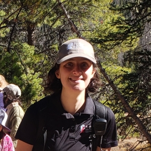 Person wearing a cap and black shirt with a walkie-talkie, smiling outdoors with trees in the background.