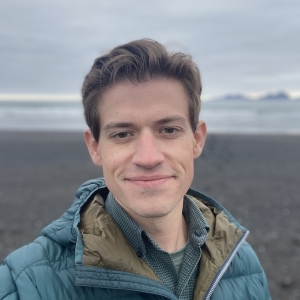 A person with brown hair wearing a teal jacket is standing on a beach with the ocean and cloudy sky in the background.