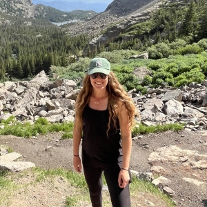 A person in a black top and pants stands on a rocky trail with a forested mountain landscape in the background, wearing a green baseball cap and sunglasses.