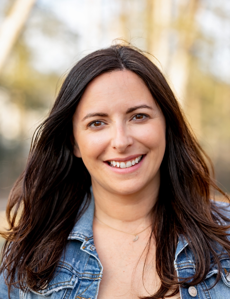 A woman with long brown hair smiles, wearing a denim jacket. The background is blurred with trees.